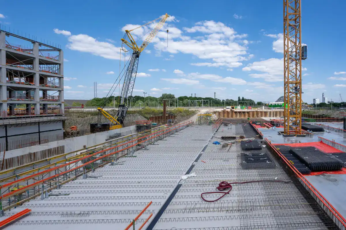 A large commercial construction site with a crawler crane working over a wide mat of reinforcing steel laid out for a structural slab.
