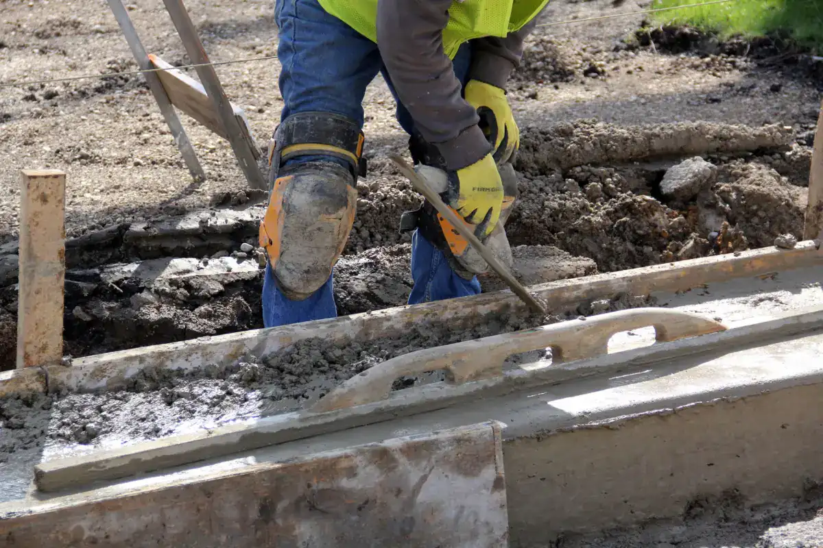 A worker in gloves and knee pads working along a formed concrete edge with a hand tool during a repair.