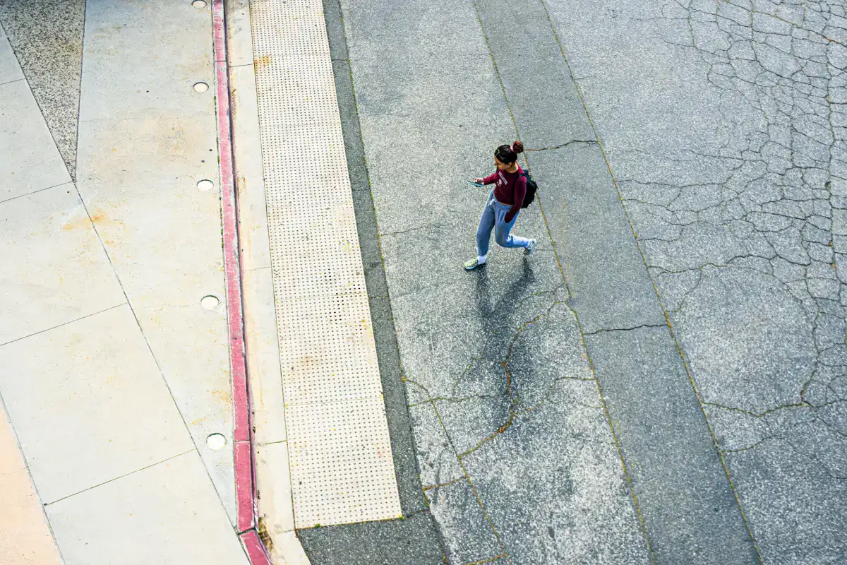 A concrete sidewalk meeting a curb ramp fitted with a detectable warning pad, with a pedestrian walking across and cracking visible in the adjoining pavement.