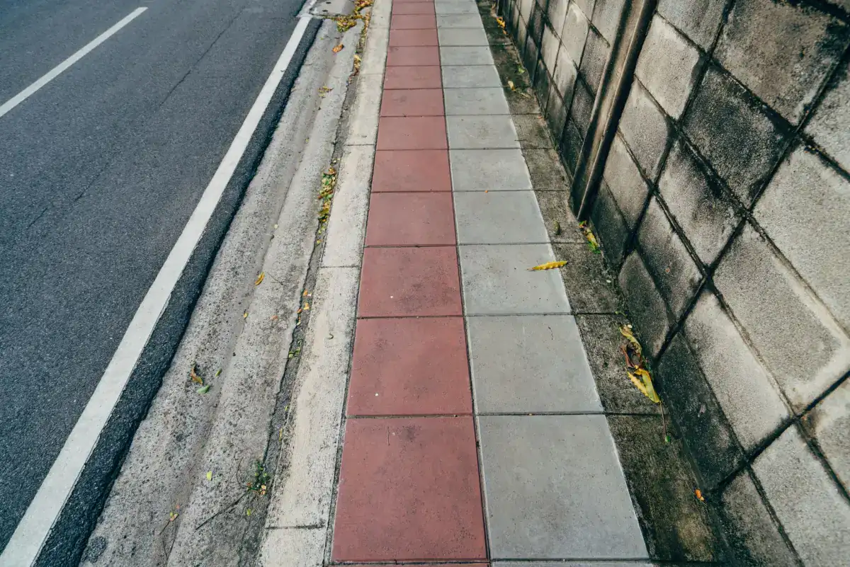 A concrete sidewalk divided into jointed squares running beside a street, with the curb line on one side and a wall on the other.
