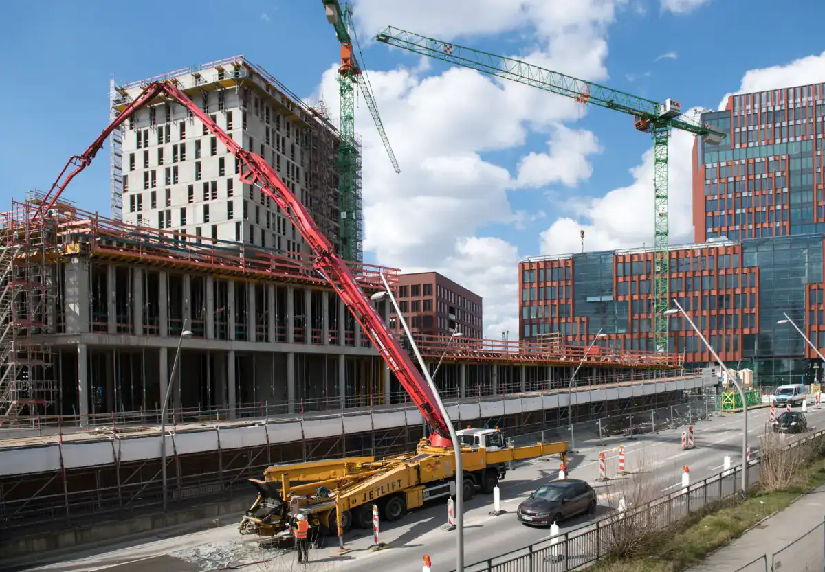 A concrete boom pump reaching over a multi-storey commercial building under construction, with tower cranes working behind it.