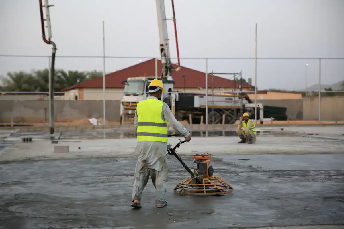 A worker in a high-visibility vest running a power trowel across a large fresh concrete slab, with a concrete pump set up behind the pour.