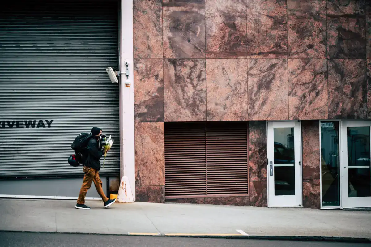 A commercial building frontage with a concrete sidewalk running past a roller shutter and glazed entrance doors, a pedestrian walking along it.