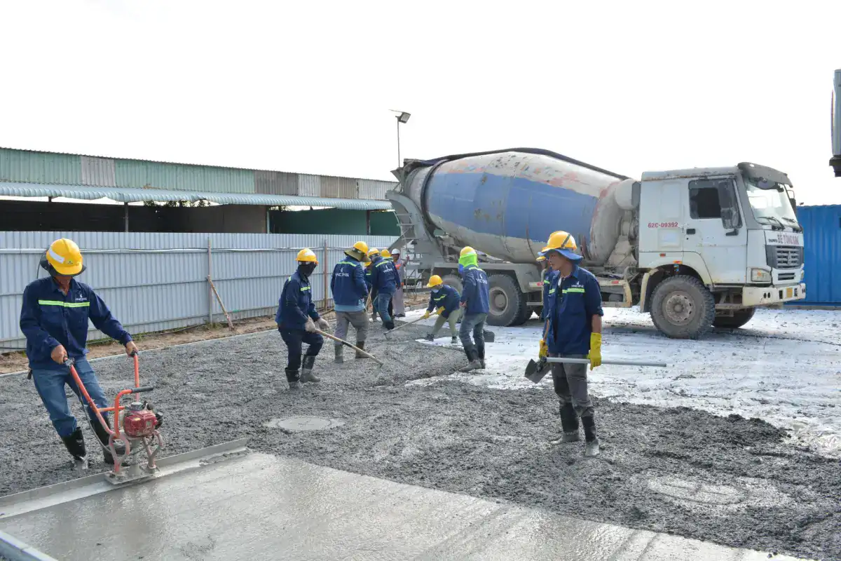 A crew in hard hats and high-visibility clothing placing and screeding fresh concrete for a slab, with a ready-mix truck discharging in the background.