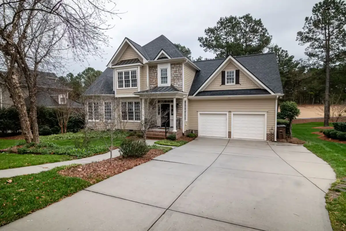A wide poured concrete driveway curving from the street to a two-car garage at a two-story suburban home, with a concrete front walk alongside and bare early-spring trees behind the house.