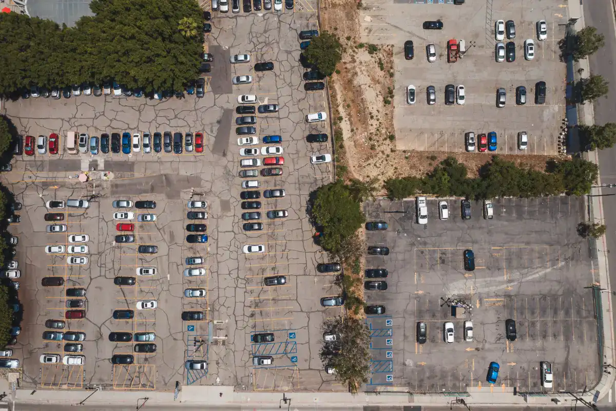 An aerial view of a large parking lot filled with cars, with cracking and patched sections visible across the pavement between the bays.