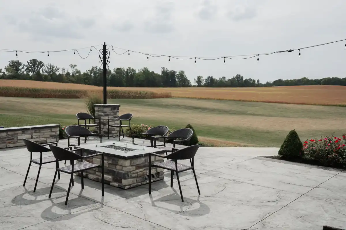 A large broom-finished concrete patio with a fire table and a ring of chairs, set at the edge of a lawn that opens onto a harvested field and a distant tree line.