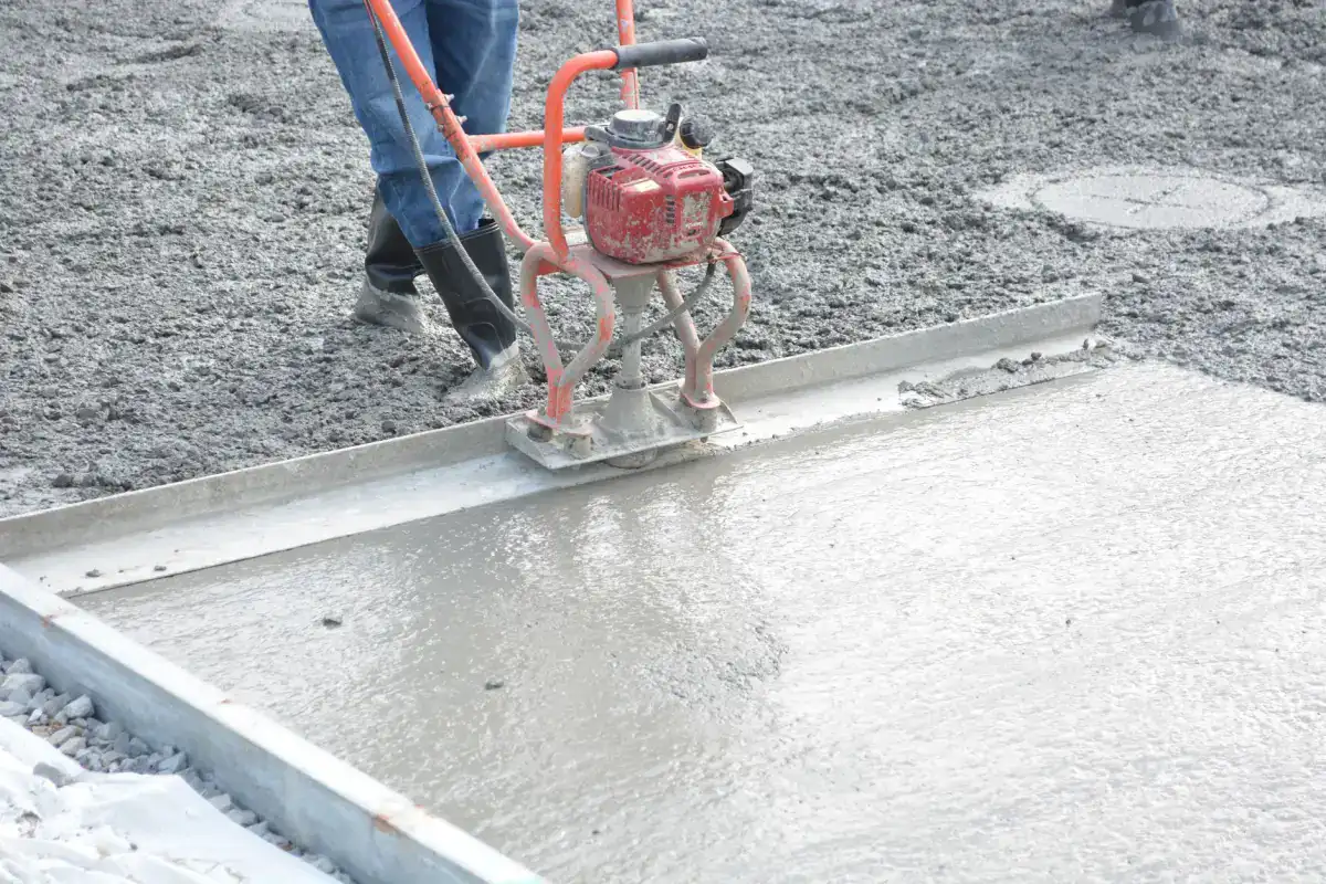 A worker running a power screed across a freshly placed concrete slab held between steel side forms, levelling the surface before finishing begins.