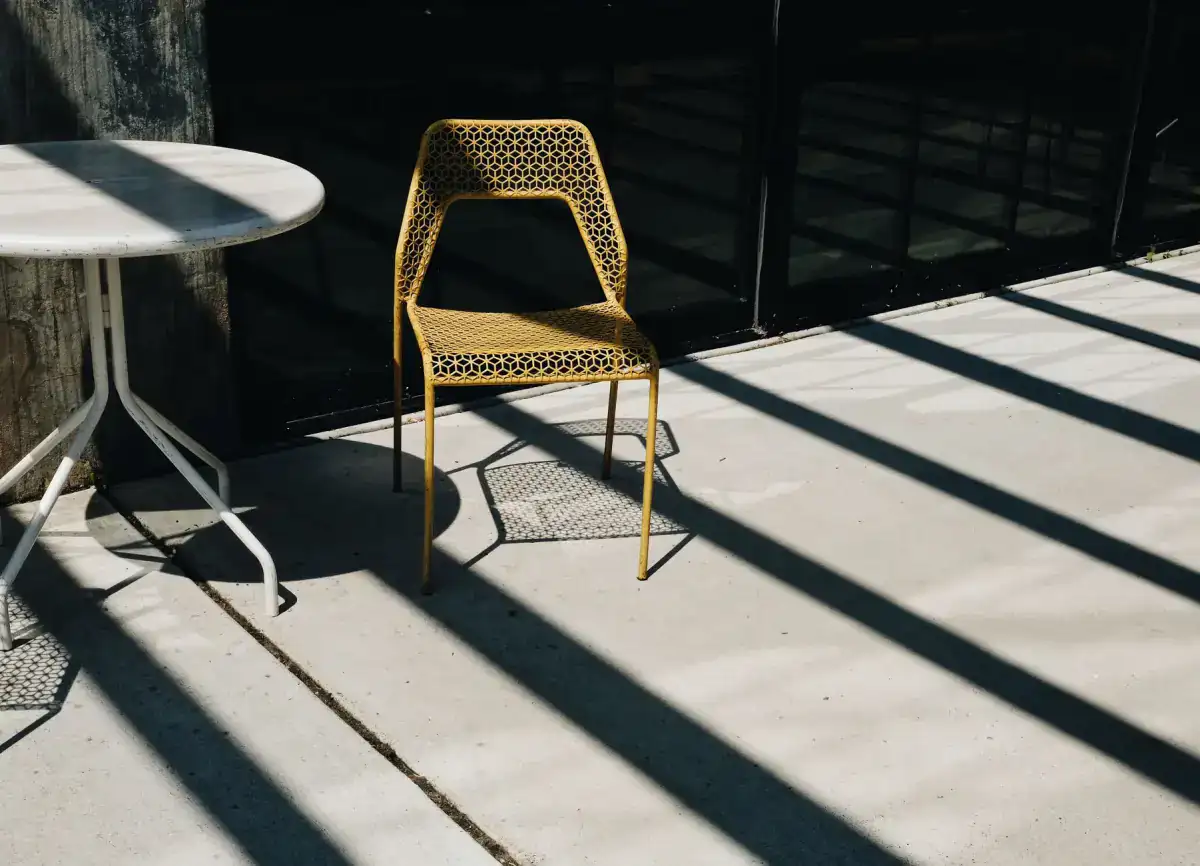 A round table and a yellow chair on a pale concrete patio, with long shadows raking across the slab and a straight sawn joint running through the frame.