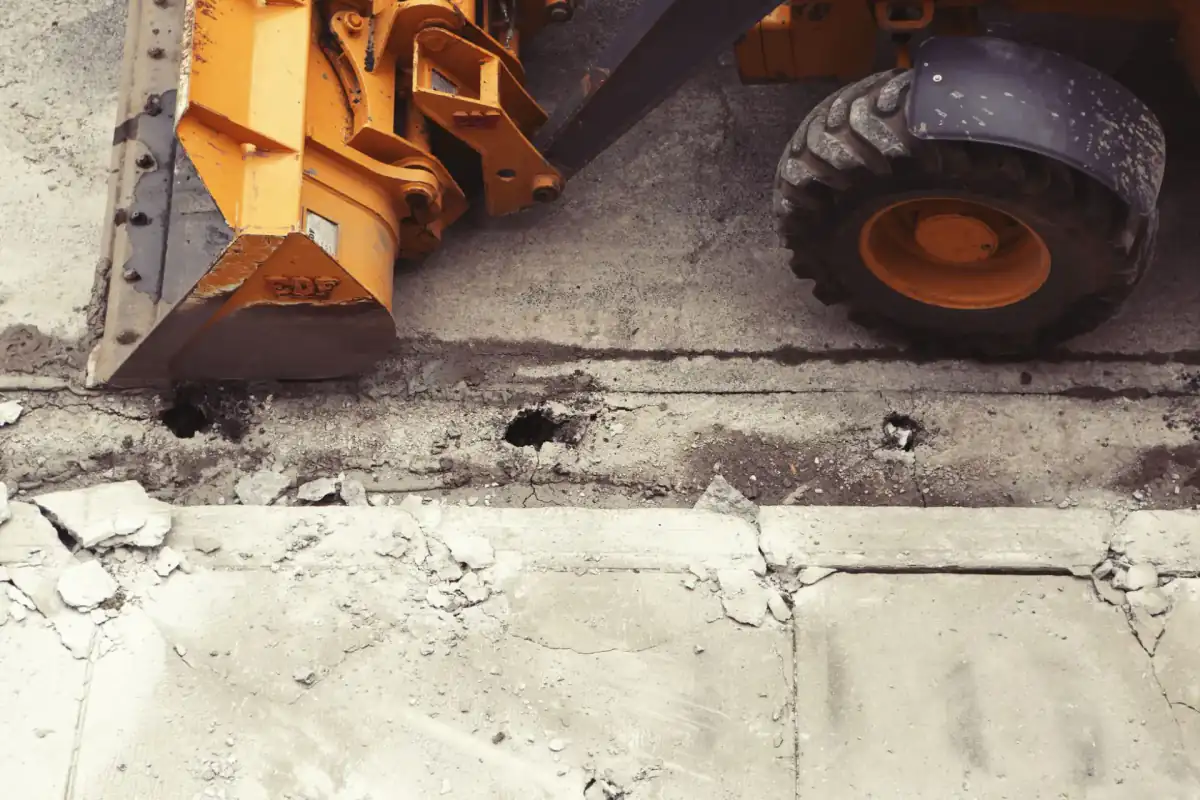 A loader bucket and wheel standing beside a broken concrete slab edge, with rubble and exposed subgrade where the pavement has been taken out.