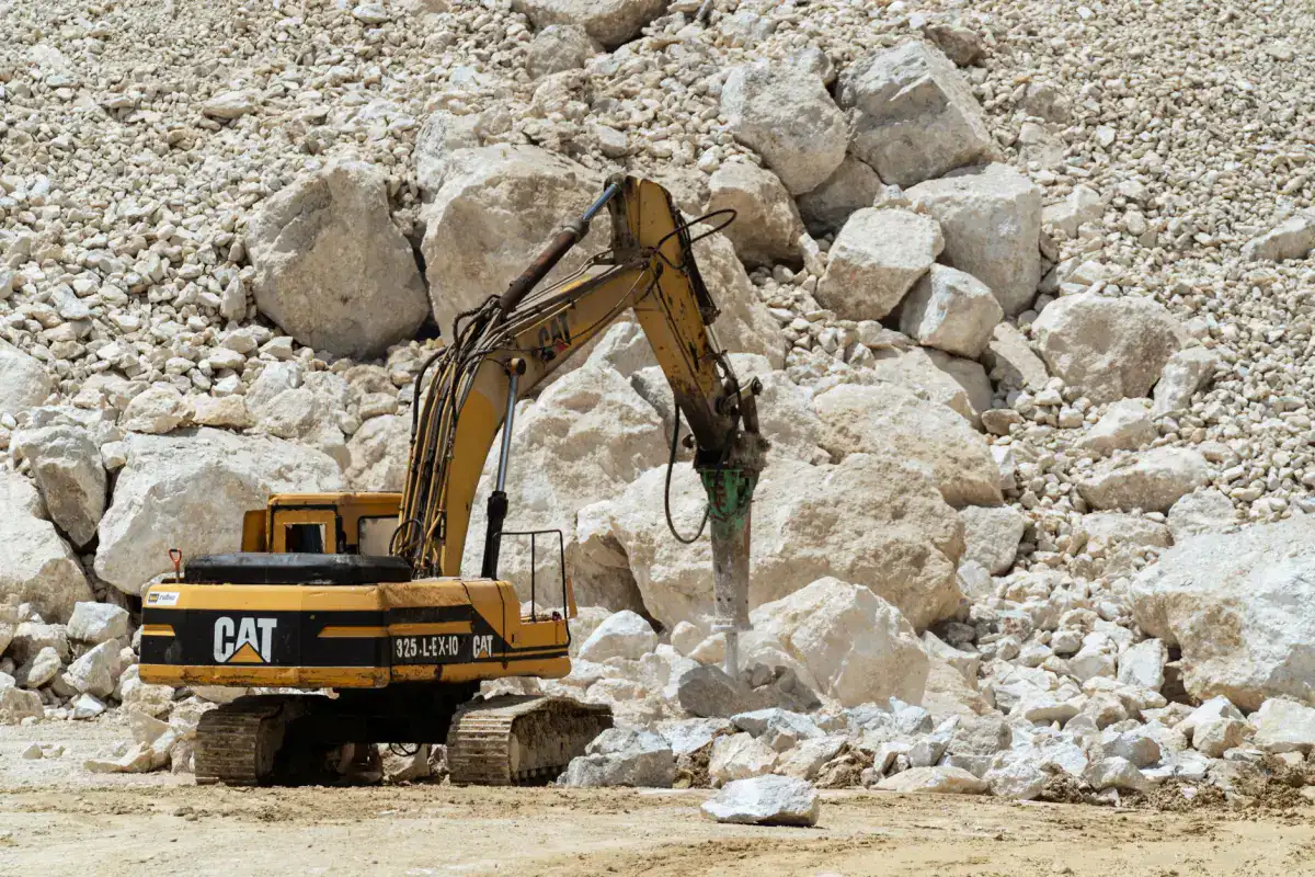 A tracked excavator fitted with a hydraulic breaker working on a large pile of broken concrete rubble.