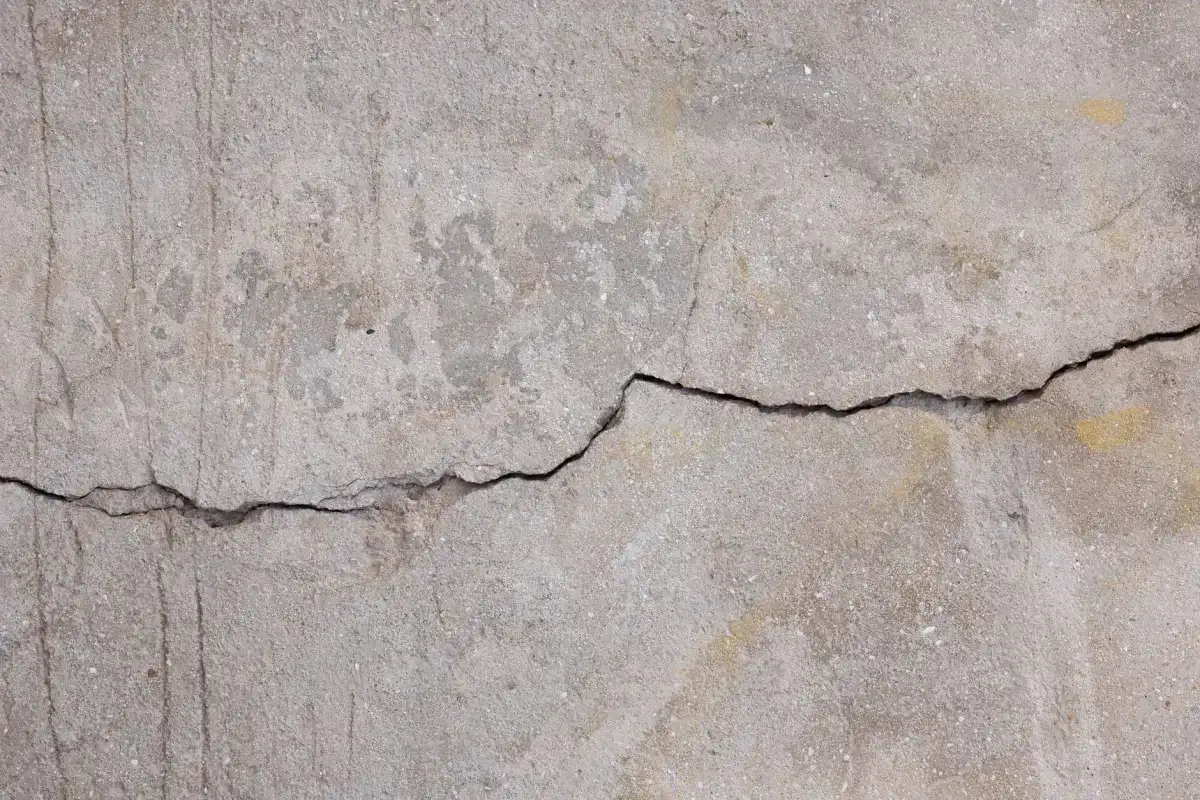 A wide, dark crack running across a weathered grey concrete surface, open enough to hold water and de-icing brine through a winter.