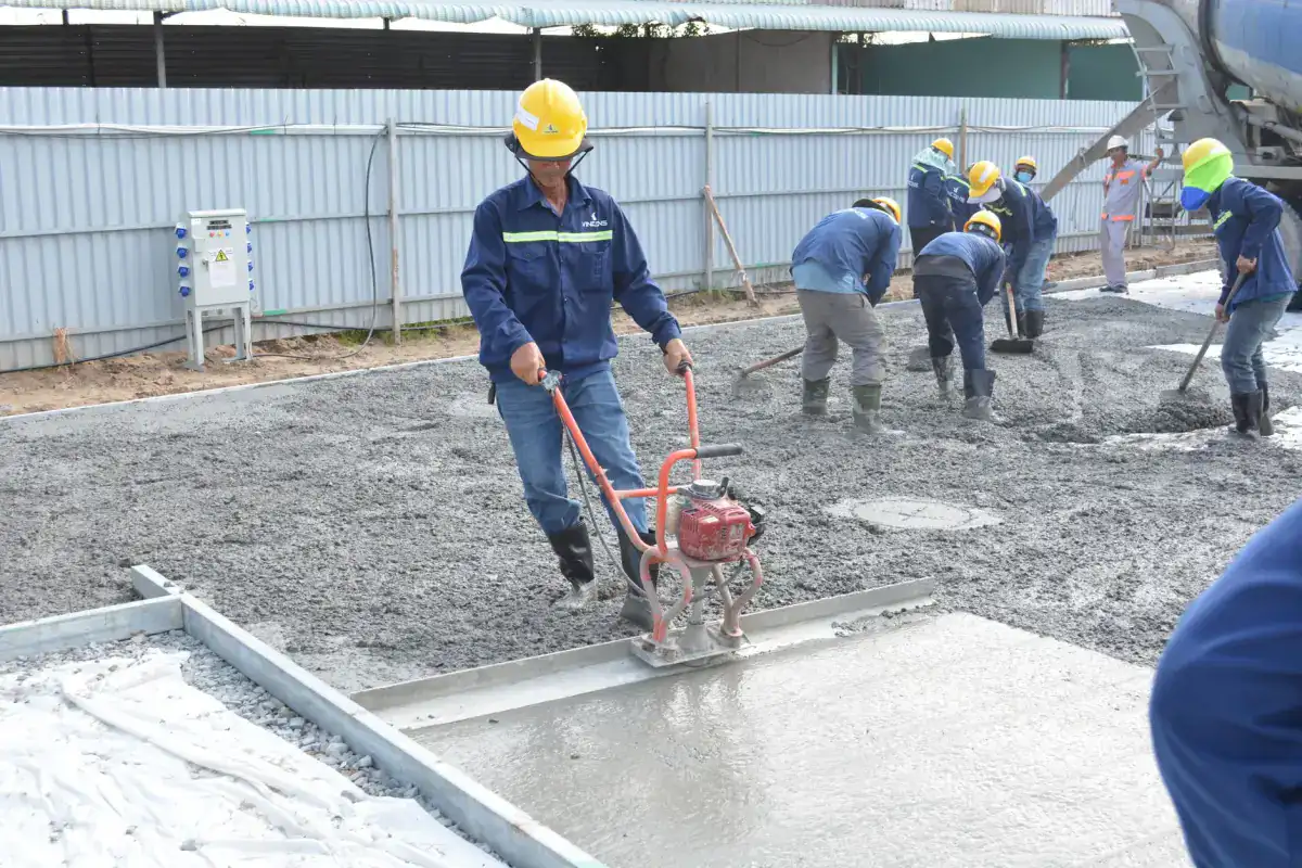 A worker guiding a power screed along a formed concrete slab while the rest of the crew places and rakes concrete behind him.