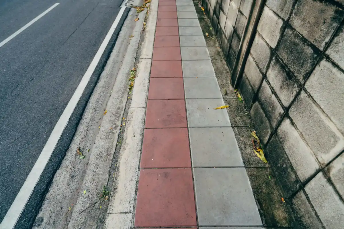 A concrete sidewalk running beside a street, divided into squares by tooled joints, with the curb and gutter line on one side and a wall on the other.
