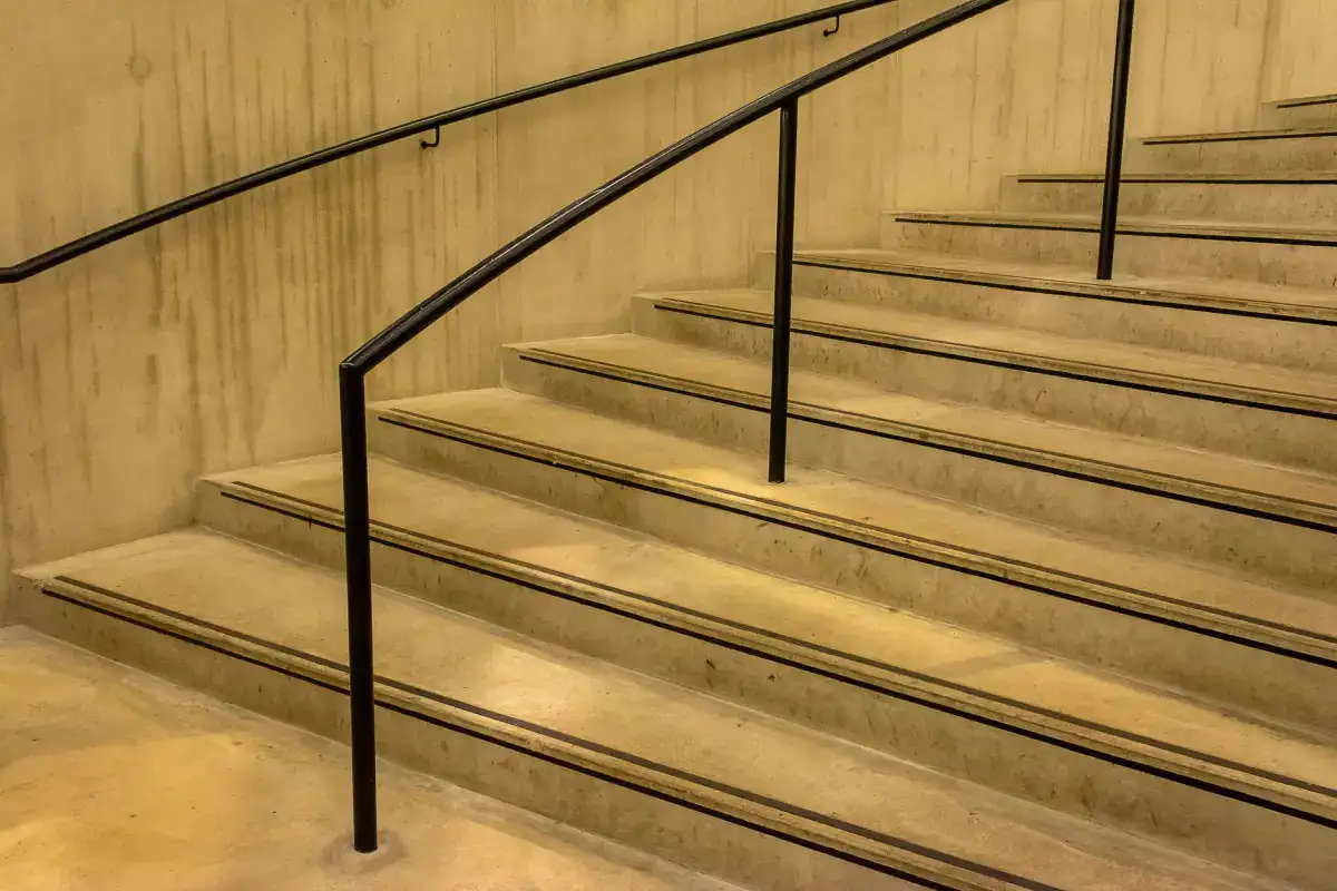 A flight of concrete steps with slim metal handrails, the treads and risers cast in consistent dimensions against a plain concrete wall.