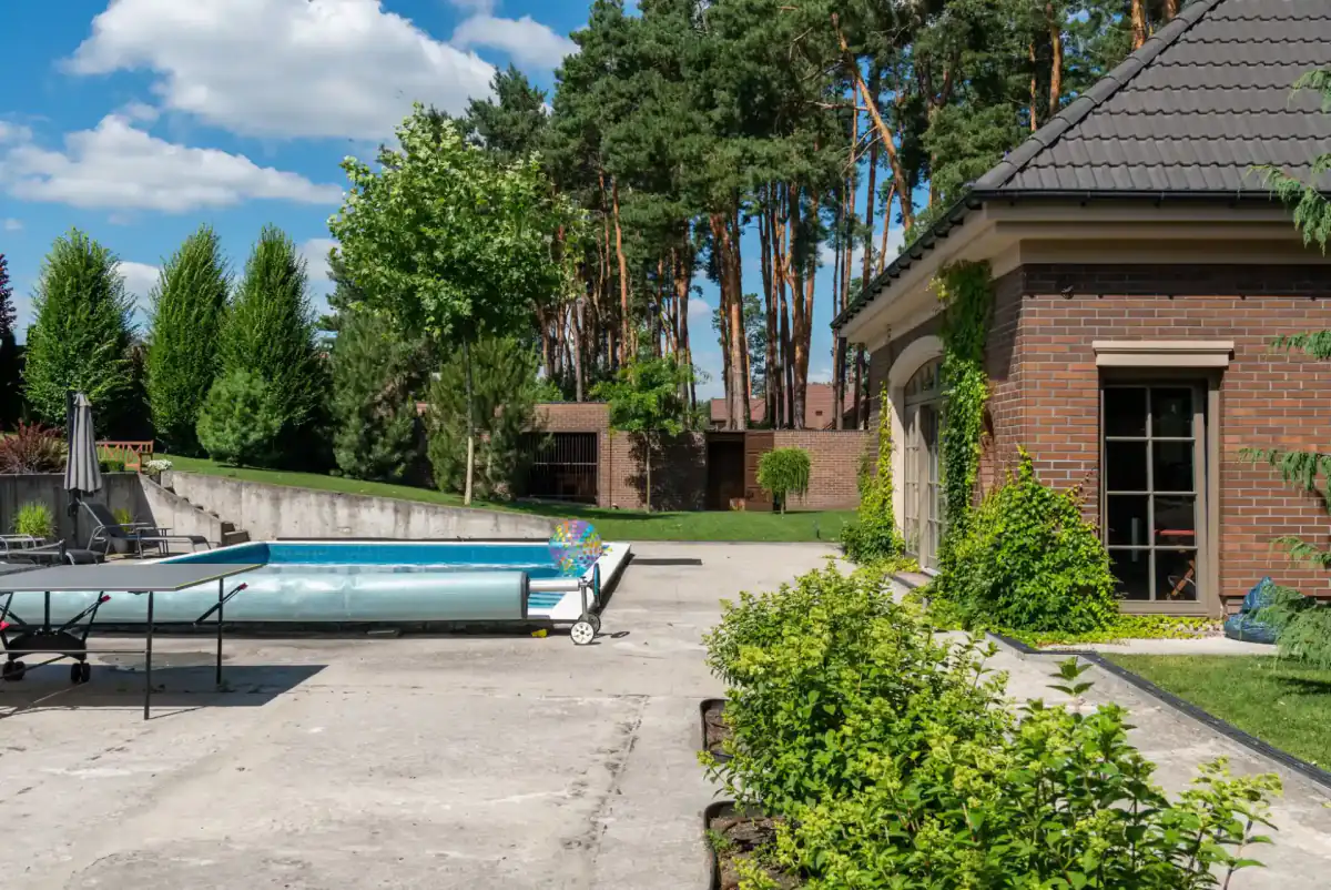 A large decorative concrete patio wrapping around a swimming pool beside a brick house, with the slab running out to shrub planting and pine trees.
